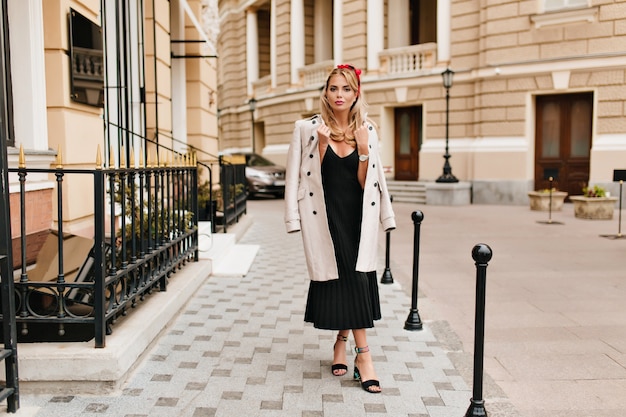 Ritratto a figura intera di bella donna in abito vintage in piedi con le gambe incrociate davanti a splendidi edifici. Foto all'aperto di una ragazza bionda affascinante che indossa un cappotto marrone chiaro e scarpe alla moda.