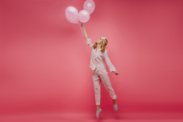 Ritratto a figura intera di bella ragazza spensierata in piedi in punta di piedi con palloncini. Foto dell'interno della signora riccia in indumenti da notte rosa e maschera per gli occhi che salta con il sorriso.