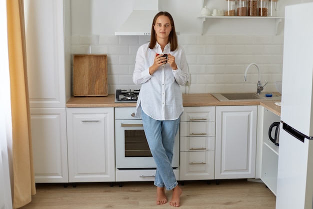 Ritratto a figura intera di donna che beve caffè al mattino a casa, in piedi vicino al set da cucina, gustando una bevanda calda a casa, indossando jeans e camicia bianca.