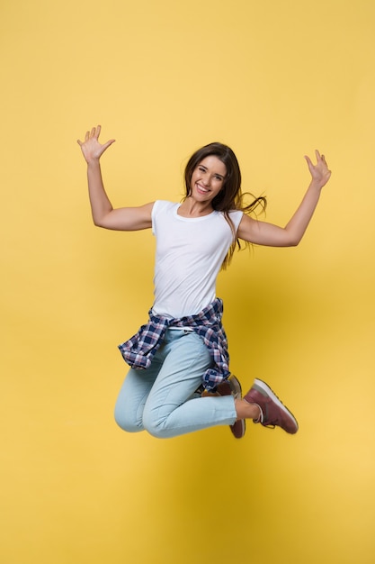 Ritratto a figura intera di ragazza spensierata in camicia bianca e jeans che saltano su sfondo giallo.