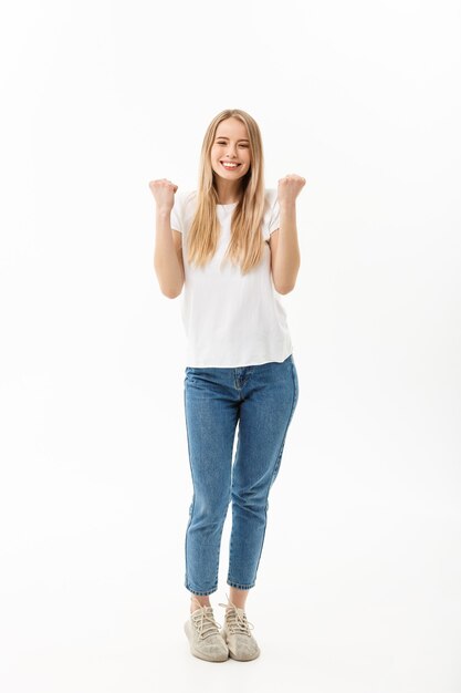 Ritratto a figura intera di una donna allegra in camicia bianca e jeans che celebra il suo successo su sfondo bianco.