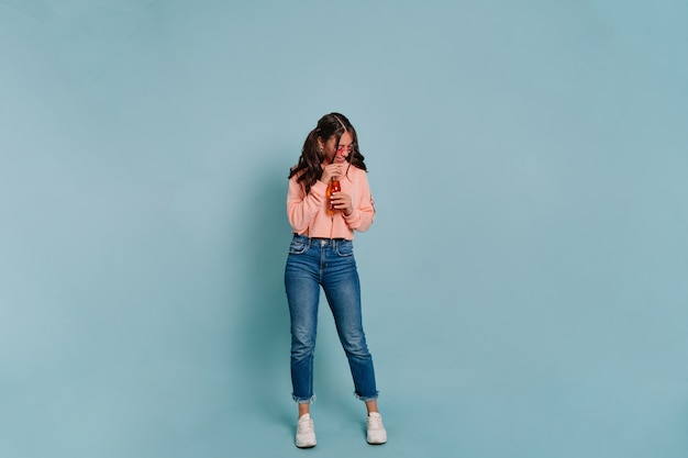 Ritratto a figura intera in studio di donna moderna ed elegante che indossa un maglione rosa e jeans in bicchieri rosa, bere succo e posa durante il servizio fotografico