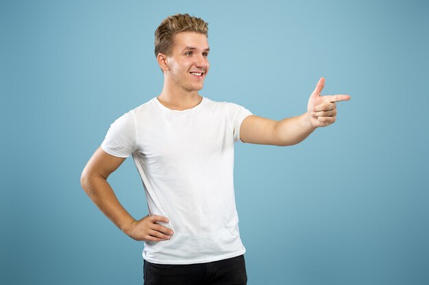 Ritratto a mezzo busto del giovane caucasico su sfondo blu studio. Bellissimo modello maschile in camicia. Concetto di emozioni umane, espressione facciale, vendite, annuncio. Indicando il lato, sorridendo.