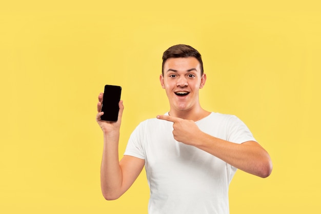 Ritratto a mezzo busto del giovane caucasico su sfondo giallo studio. Bellissimo modello maschile in camicia. Concetto di emozioni umane, espressione facciale, vendite, annuncio. Mostrando lo schermo del telefono e sorridendo.