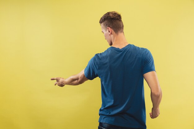 Ritratto a mezzo busto del giovane caucasico su sfondo giallo studio. Bello modello maschio in camicia blu. Concetto di emozioni umane, espressione facciale. Mostrare e indicare qualcosa. Invitante.