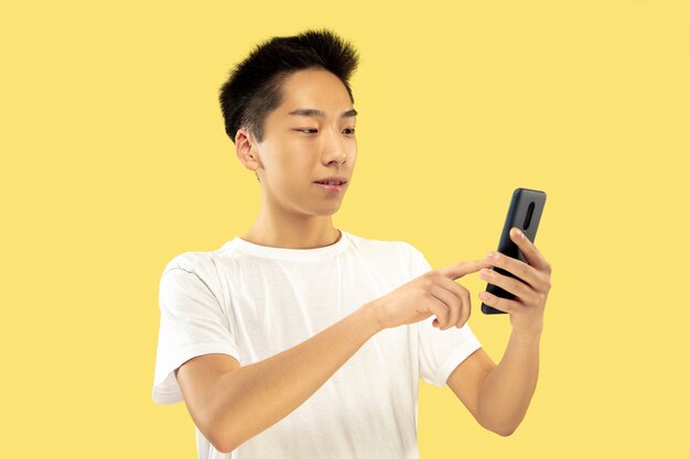 Ritratto a mezzo busto del giovane coreano su sfondo giallo studio. Modello maschile in camicia bianca. Utilizzando smartphone. Concetto di emozioni umane, espressione facciale. Vista frontale. Colori alla moda.