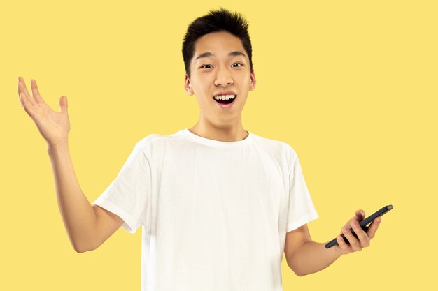 Ritratto a mezzo busto del giovane coreano su sfondo giallo studio. Modello maschile in camicia bianca. Utilizzo dello smartphone per scommettere, leggere notizie o parlare. Concetto di emozioni umane, espressione facciale.