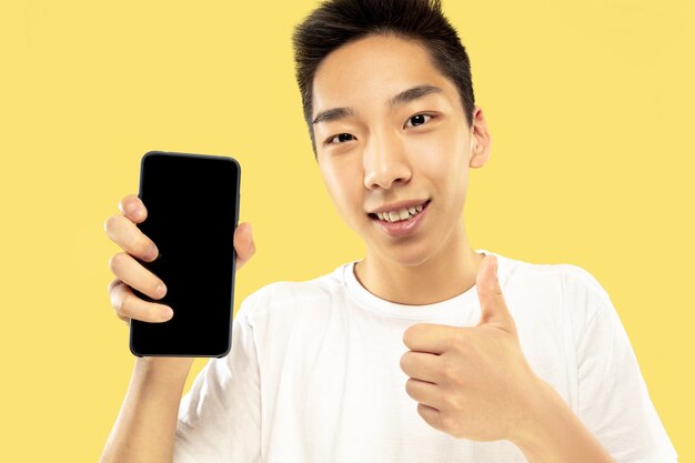 Ritratto a mezzo busto del giovane coreano su sfondo giallo studio. Modello maschile in camicia bianca. Utilizzo dello smartphone per scommettere, leggere notizie o parlare. Concetto di emozioni umane, espressione facciale.