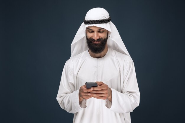 Ritratto a mezzo busto dell'uomo arabo saudita su sfondo blu scuro per studio. Giovane modello maschio utilizza lo smartphone, in chat. Concetto di affari, finanza, espressione facciale, emozioni umane, tecnologie.