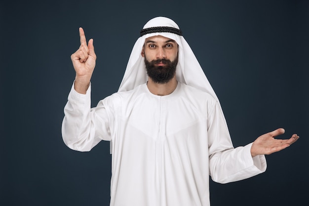 Ritratto a mezzo busto dell'uomo arabo saudita sulla parete blu scuro. Giovane modello maschio che sorride e che indica. Concetto di affari, finanza, espressione facciale, emozioni umane, tecnologie.