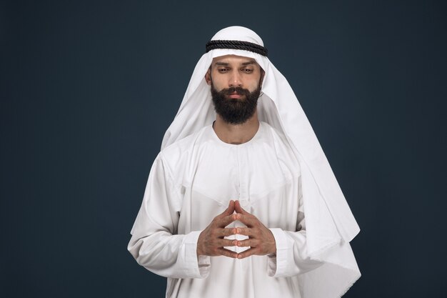 Ritratto a mezzo busto dell'uomo d'affari arabo saudita su sfondo blu scuro per studio. Giovane modello maschio che prega e sembra premuroso. Concetto di affari, finanza, espressione facciale, emozioni umane.