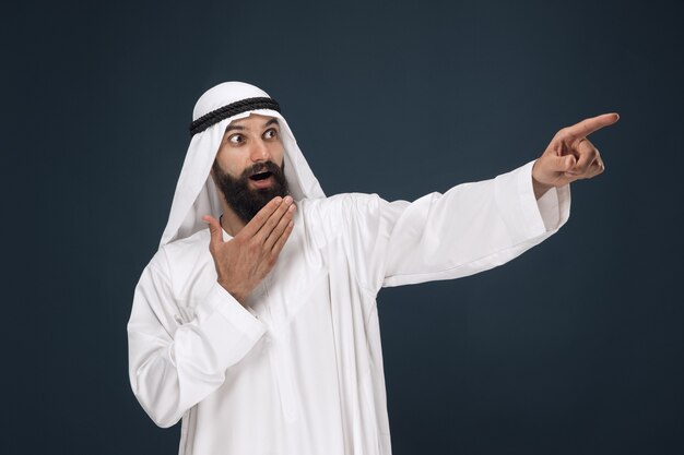 Ritratto a mezzo busto dell'uomo d'affari arabo saudita su sfondo blu scuro per studio. Giovane modello maschio stupito, indicando o scegliendo. Concetto di affari, finanza, espressione facciale, emozioni umane.