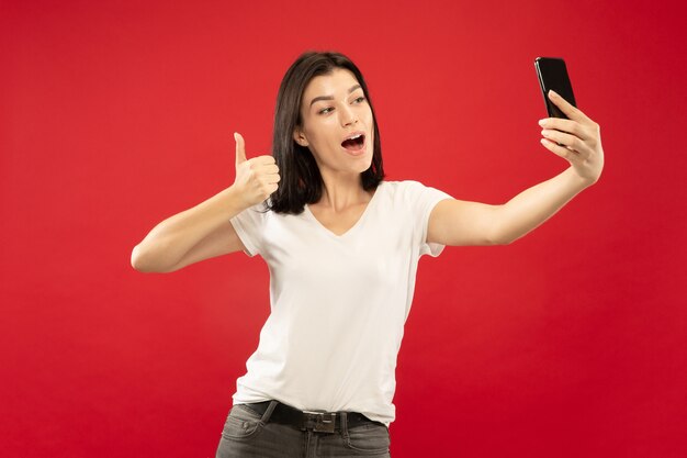 Ritratto a mezzo busto della giovane donna caucasica su sfondo rosso studio. Bello modello femminile in camicia bianca. Concetto di emozioni umane, espressione facciale. Fare selfie o il proprio vlog, blog.