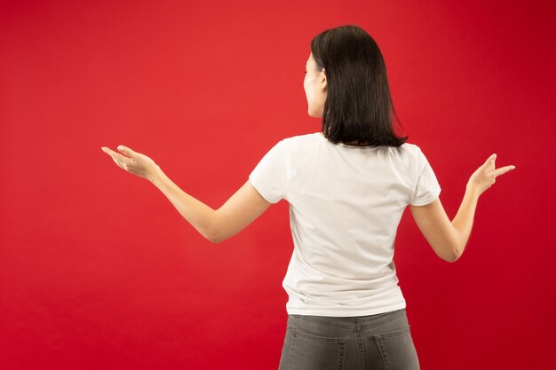 Ritratto a mezzo busto della giovane donna caucasica su sfondo rosso studio. Bello modello femminile in camicia bianca. Concetto di emozioni umane, espressione facciale. Mostrare e indicare qualcosa.