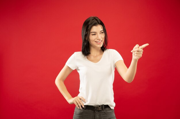 Ritratto a mezzo busto della giovane donna caucasica su sfondo rosso studio. Bello modello femminile in camicia bianca. Concetto di emozioni umane, espressione facciale. Mostrare, indicare qualcosa, sembra giocoso.