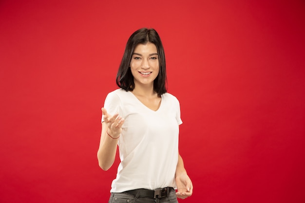 Ritratto a mezzo busto della giovane donna caucasica su sfondo rosso studio. Bello modello femminile in camicia bianca. Concetto di emozioni umane, espressione facciale. Mostrare qualcosa, invitare, scegliere.