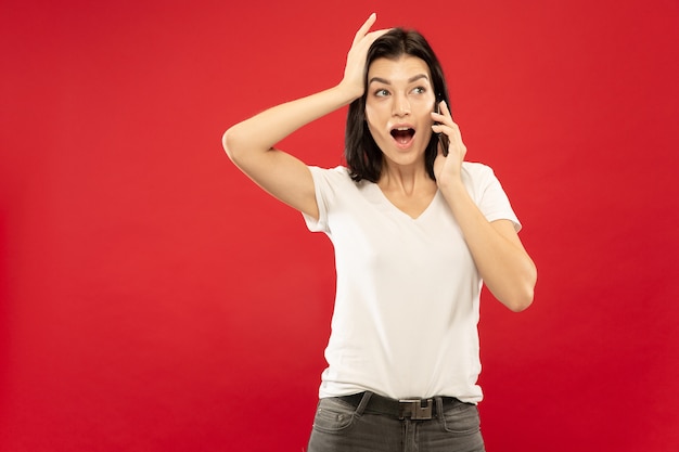 Ritratto a mezzo busto della giovane donna caucasica su sfondo rosso studio. Bello modello femminile in camicia bianca. Concetto di emozioni umane, espressione facciale. Parlando su smartphone, espressivo.