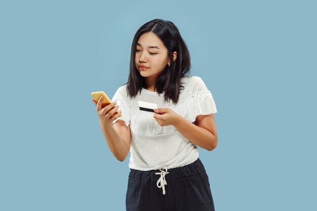 Ritratto a mezzo busto della giovane donna coreana su spazio blu. Modello femminile che utilizza il suo smartphone per pagare la bolletta o acquisti online.