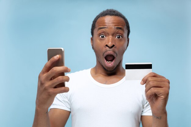 Ritratto a mezzo busto di giovane uomo afro-americano in camicia bianca che tiene una carta e smartphone su spazio blu