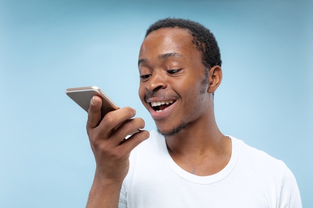 Ritratto a mezzo busto di giovane uomo afro-americano in camicia bianca su sfondo blu. Emozioni umane, espressione facciale, annuncio, concetto di vendita. Tenere in mano uno smartphone, parlare o registrare un messaggio vocale.