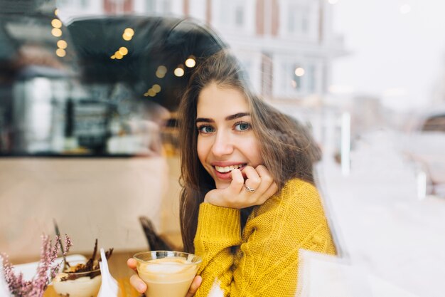 Ritratto affascinante giovane donna con un sorriso amichevole, lunghi capelli castani sorridente nella finestra del caffè nel periodo invernale. Vere emozioni positive, tempo libero, bere caffè, rilassarsi quando fa freddo.