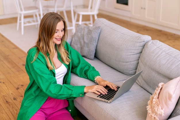 Ritratto al coperto di una donna bionda bionda freelance che lavora sul suo laptop al concetto di ufficio a casa del soggiorno indossando camicia e pantaloni luminosi