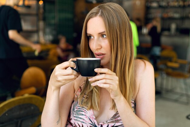 Ritratto al coperto o bella donna bionda che beve un gustoso cappuccino al ristorante, godetevi la sua colazione, un vestito elegante, un trucco naturale.