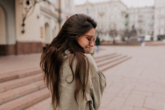 Ritratto all'aperto dal retro della giovane donna alla moda con giacca vestita di capelli lunghi che gode del tempo fuori