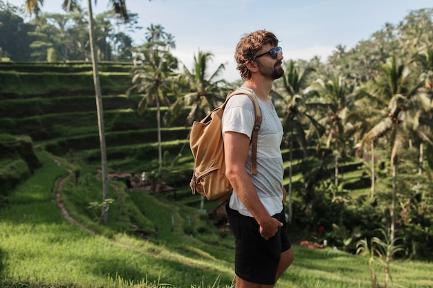 Ritratto all'aperto dell'uomo bello di viaggio con lo zaino che cammina sulla terrazza di aumento a Bali.