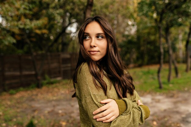 Ritratto all'aperto di affascinante donna abbastanza europea con lunghi capelli ondulati che indossa un maglione verde che cammina nel parco