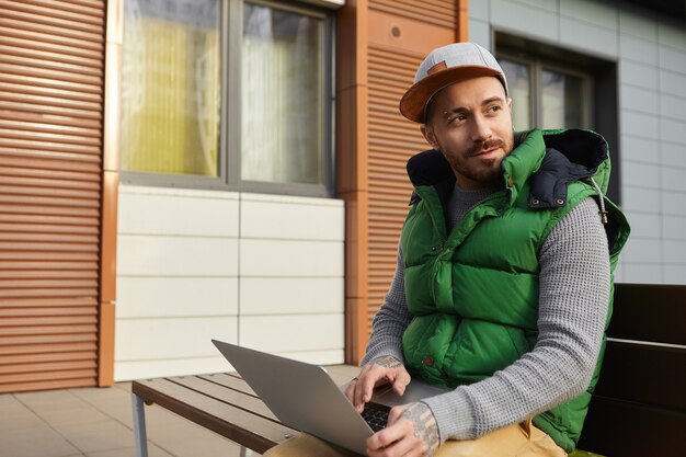 Ritratto all'aperto di elegante freelance maschio con la barba lunga in abiti alla moda seduto su una panchina con il computer portatile sulle ginocchia, utilizzando la connessione Internet wireless 4g per il lavoro online, la tastiera