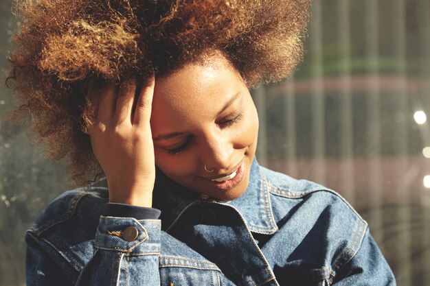 Ritratto all'aperto di giovane donna dalla carnagione scura alla moda con capelli ricci che indossa giacca di jeans