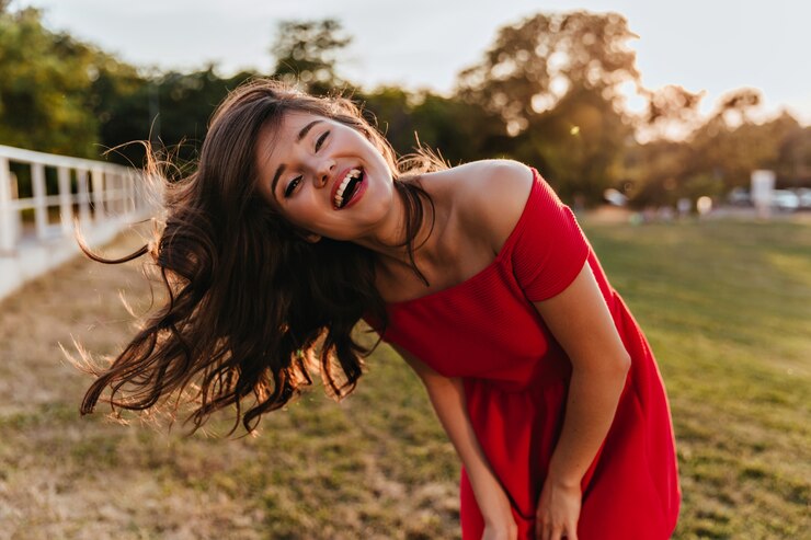 Foto gratuito ritratto all'aperto di giovane signora di buon umore in vestito rosso che esprime felicità. foto della ragazza soddisfatta con capelli castani che fluttuano in posa sulla natura
