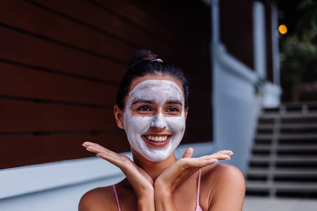 Ritratto all'aperto di pelle abbronzata calma donna abbastanza caucasica in bikini al centro benessere con maschera peeling bianca sul viso