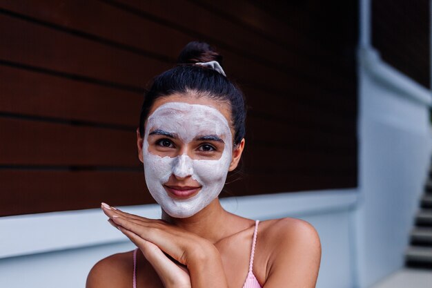 Ritratto all'aperto di pelle abbronzata calma donna abbastanza caucasica in bikini al centro benessere con maschera peeling bianca sul viso