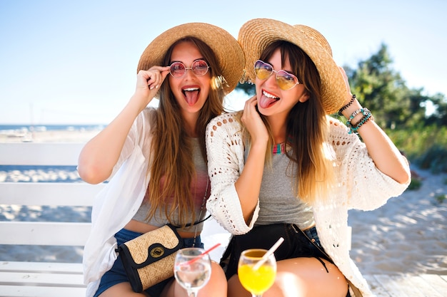 Ritratto all'aperto di ragazze hipster divertenti e felici della compagnia che impazziscono sul caffè sulla spiaggia, bevendo gustosi cocktail ridendo e sorridendo, abiti estivi vintage boho luminosi, relazioni e divertimento.