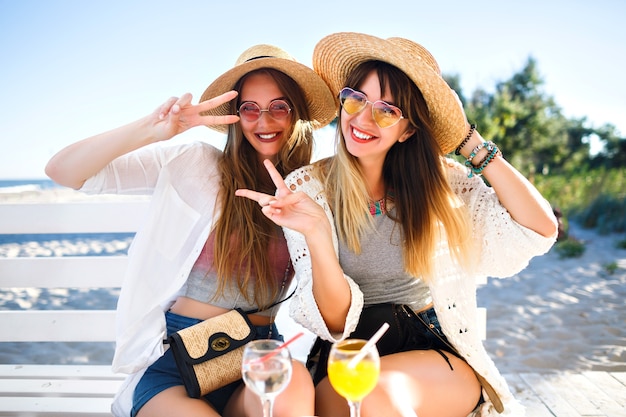 Ritratto all'aperto di ragazze hipster divertenti e felici della compagnia che impazziscono sul caffè sulla spiaggia, bevendo gustosi cocktail ridendo e sorridendo, abiti estivi vintage boho luminosi, relazioni e divertimento.