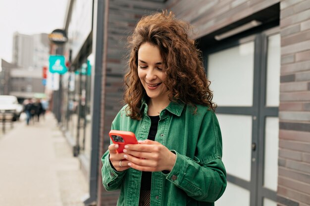 Ritratto all'aperto di una bella ragazza alla moda con riccioli che indossa una camicia verde utilizzando lo smartphone con il sorriso La giovane ragazza caucasica spensierata utilizza uno smartphone moderno in piedi all'aperto