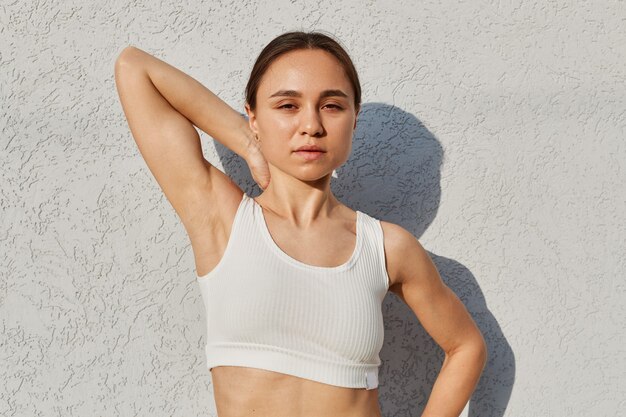 Ritratto all'aperto di una giovane ragazza atletica in posa vicino al muro bianco con il braccio alzato, guardando la telecamera, fotografata durante l'allenamento, stile di vita sano.