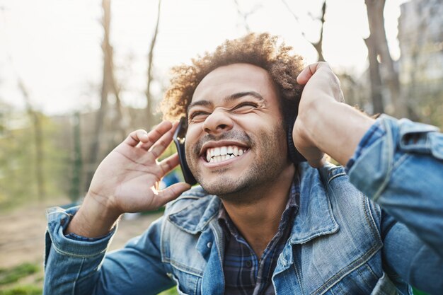 Ritratto all'aperto di vista laterale dell'uomo africano felice eccitato con l'acconciatura afro che tiene le cuffie mentre ascolta la musica e sorride ampiamente, stupito da ciò che sente.