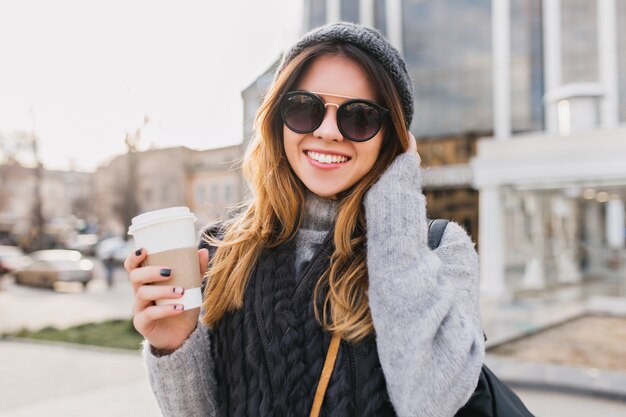 Ritratto alla moda urbana giovane donna con caffè per andare a piedi nel soleggiato centro città. Incredibile donna sorridente in occhiali da sole moderni, cappello lavorato a maglia, maglione di lana divertendosi all'aperto.
