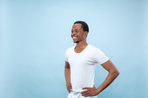 Ritratto alto vicino a mezzo busto di giovane uomo afro-americano in camicia bianca su sfondo blu. Emozioni umane, espressione facciale, concetto di annuncio. In piedi e sorridente.