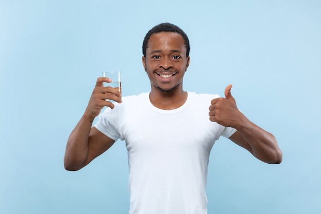 Ritratto alto vicino a mezzo busto di giovane uomo afro-americano in camicia bianca su sfondo blu. Emozioni umane, espressione facciale, concetto di annuncio. Tenendo un bicchiere e acqua potabile.