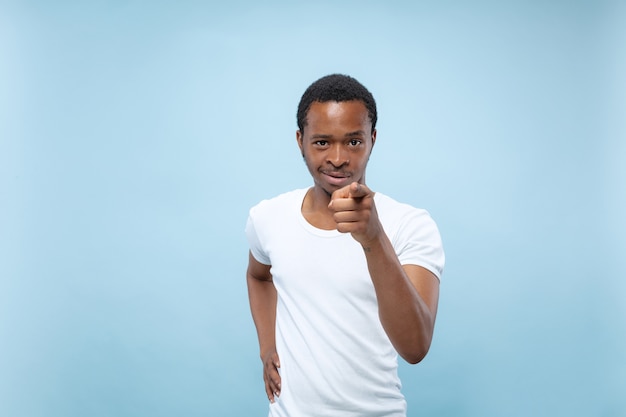 Ritratto alto vicino a mezzo busto di giovane uomo afro-americano in camicia bianca su spazio blu. Emozioni umane, espressione facciale, concetto di annuncio. Indicare, scegliere e sorridere