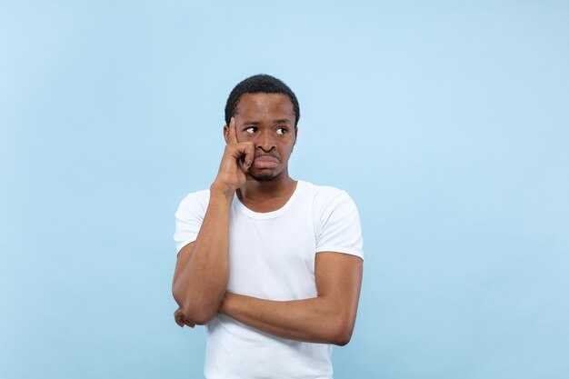 Ritratto alto vicino a mezzo busto di giovane uomo afro-americano in camicia bianca sulla parete blu. Emozioni umane, espressione facciale, concetto di annuncio. Premuroso, pensando che copre il viso con le mani.