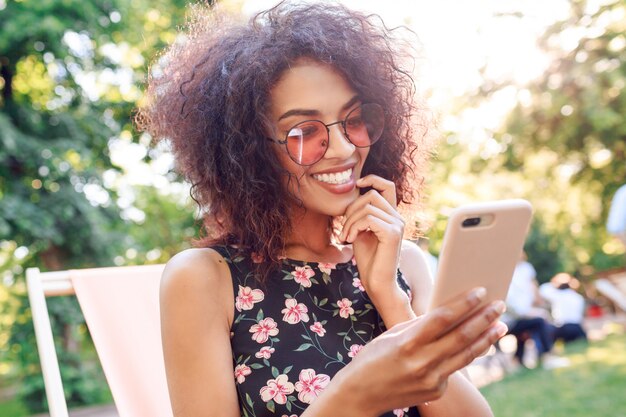 Ritratto alto vicino all'aperto della donna di colore sorridente che per mezzo del telefono cellulare e facendo autoritratto