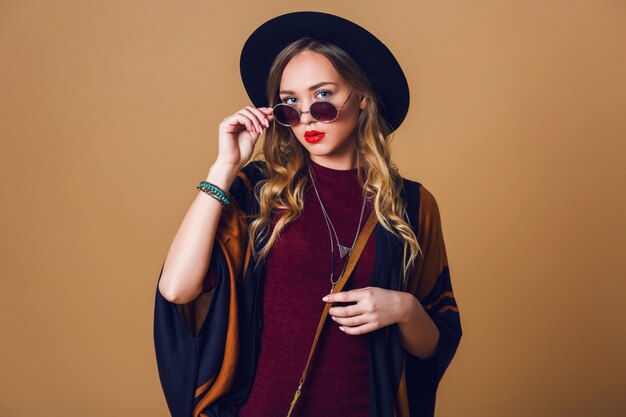 Ritratto alto vicino dello studio di giovane donna bionda fresca in poncio marrone della paglia, cappello d'avanguardia nero della lana e vetri rotondi che esaminano macchina fotografica. Borsa in pelle verde.