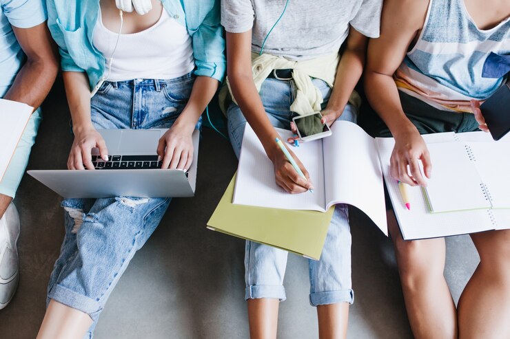 Ritratto ambientale del primo piano della ragazza in camicia blu e jeans che tengono il computer portatile sulle ginocchia mentre sedendosi accanto ai compagni dell'università. Studentessa lezione di scrittura in notebook e utilizzo del telefono tra amici