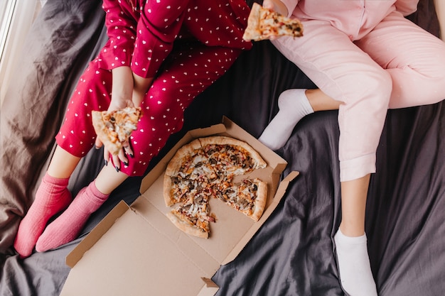 Ritratto ambientale di due ragazze in pigiama seduto sul letto con fast food italiano. Modelli femminili pigri che mangiano pizza sul foglio scuro.