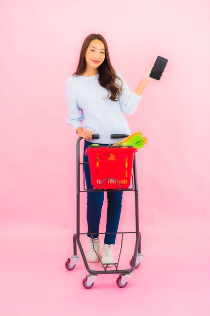 Ritratto bella giovane donna asiatica con frutta verdura e merce nel carrello della drogheria sulla parete isolata rosa